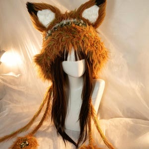 May include: A fluffy, orange and brown faux fur animal hat featuring large, upright ears with white centres and black tips. The hat includes long, braided side tassels with pom-poms at the ends, worn on a mannequin head with long, straight brown hair.