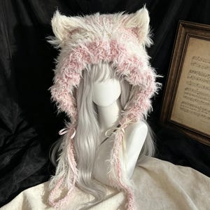 May include: A plush, hooded hat featuring cat ears, crafted from white and soft pink faux fur. The hood has a textured, shaggy appearance with long, dangling side panels and thin, light-colored ribbon ties hanging down from the front opening.