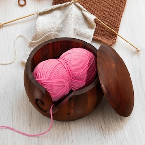 May include: A wooden yarn bowl with a lid, holding two pink yarn balls. The bowl is dark brown with a swirl design for the yarn to feed through. Knitting needles and partially knitted projects are in the background.