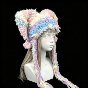 May include: A multi-colored, fuzzy, crocheted jester-style hat featuring two pointed peaks with small pom-poms at the ends. The hat is crafted from textured yarn in shades of pastel pink, blue, cream, and yellow, with long, dangling braided fringe.
