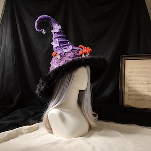 May include: A purple crocheted pointed hat with a black faux fur brim, decorated with small red crocheted mushrooms, translucent light purple butterfly appliqués, and a single clear crystal bead dangling from the curled tip of the hat.