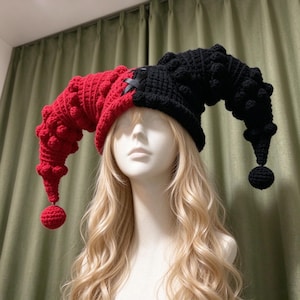 May include: A two-toned crochet jester hat featuring a red left side and a black right side. The hat has two long, curved horns with textured bobble stitches and a round pom-pom at each tip. A black ribbon is laced through the centre seam.