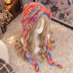 May include: A vibrant, multi-coloured crochet bonnet featuring a textured, chunky knit pattern with a blend of bright pink, orange, yellow, blue, and purple yarn. The headwear includes two long, dangling braided ties that hang down from the sides.