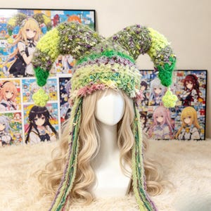 May include: A multi-textured crochet jester hat in shades of lime green, olive, and lavender. The hat features two long, curved horns topped with small, light green star-shaped charms and long, dangling yarn tassels hanging from the sides.