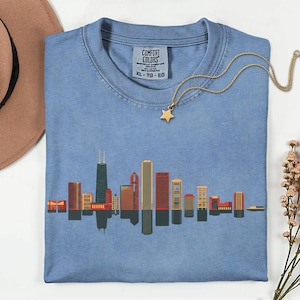 Op de afbeelding: Lichtblauw Comfort Colors XL T-shirt met een skyline-afbeelding. De afbeelding toont gebouwen in rood, oranje en geel. Een gouden ketting met een sterhanger ligt op het T-shirt. Het shirt is van katoen.