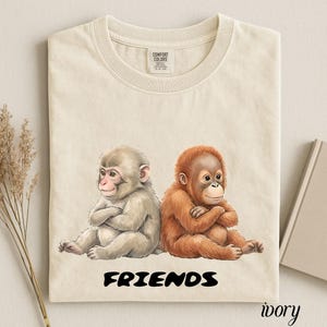 Könnte beinhalten: Elfenbeinfarbenes T-Shirt mit einer Grafik von zwei Comic-Affen, die nebeneinander mit verschränkten Armen sitzen. Das Wort "FRIENDS" ist in Schwarz unter den Affen aufgedruckt. Das Shirt ist mit "ivory" in Schrift beschriftet.