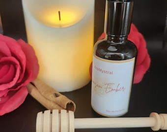 May include: A dark brown perfume bottle with a white label that reads "AroMystral pro Ember Eau de Parfum" sits next to a large cream-colored candle, a red rose, a cinnamon stick, and a wooden honey dipper.