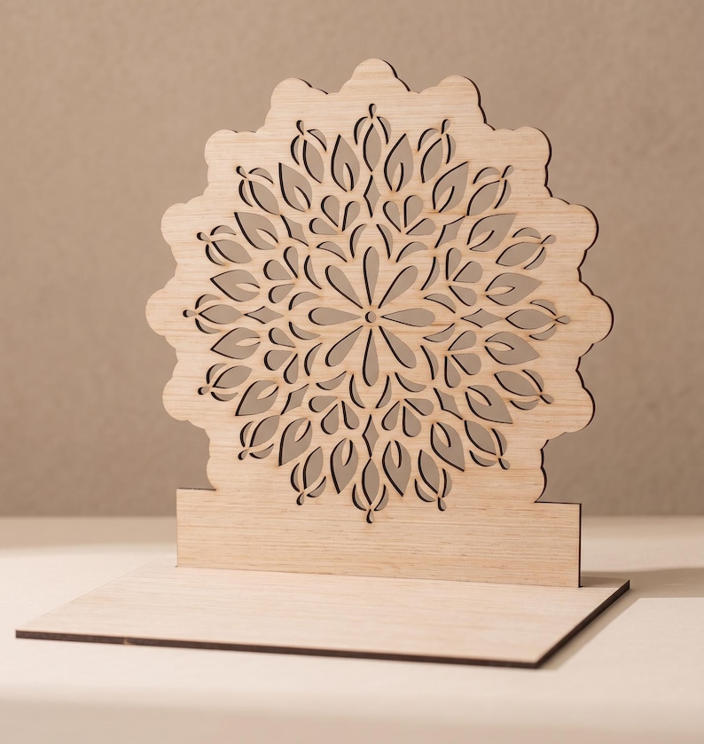 Op de afbeelding: Een lichtgekleurde houten displaystandaard met een cirkelvormig, ingewikkeld lasergesneden mandalapatroon. Het ontwerp bestaat uit symmetrische, bloembladachtige uitsparingen die vanuit een centraal punt stralen, ondersteund door een voet.