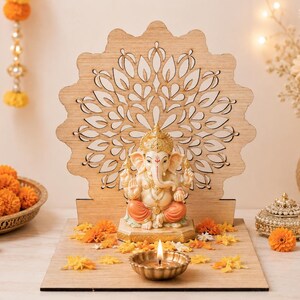 Op de afbeelding: Een lichtgekleurde houten achtergrond met een lasergesneden mandalapatroon achter een klein, zittend Ganesha-beeldje. Het beeldje heeft een gouden kroon, een oranje dhoti en een cr&egrave;mekleurige huid, rustend op een houten voet met een kleine koperen diya.