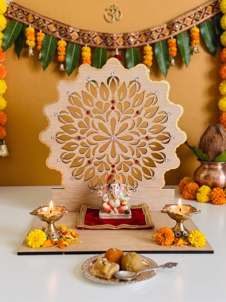 Op de afbeelding: Een houten altaar met een lasergesneden mandala-achtergrond met ingebedde kristallen, een klein Ganesha-beeldje op een rood fluwelen kussen, twee brandende koperen olielampen en een bord met snoepjes op een platte houten basis.