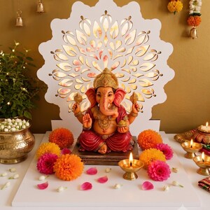 Op de afbeelding: Een standbeeld van de godheid Ganesha met een roze huid en rode gewaden, zittend op een vierkante voet voor een witte, lasergesneden, cirkelvormige decoratieve achtergrond. De sc&egrave;ne is versierd met oranje en roze goudsbloemen en koperen olielampen.