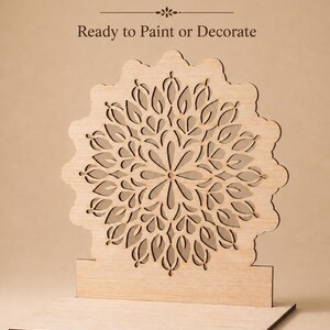 Op de afbeelding: Een lichtgekleurde houten uitsparing met een cirkelvormig mandalapatroon met ingewikkelde bloembladachtige openingen, gemonteerd op een rechthoekige voet. De tekst "DIY Unfinished Product" en "Ready to Paint or Decorate" staat boven het object.