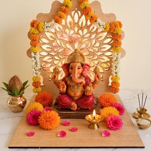 Op de afbeelding: Een Ganesha-beeld met een roze huid en rode kleding staat op een houten voetstuk voor een lasergesneden houten achtergrond. Het geheel is versierd met oranje en roze afrikaantjes, een koperen olielamp, een kokosnoot in een koperen pot en wierook.