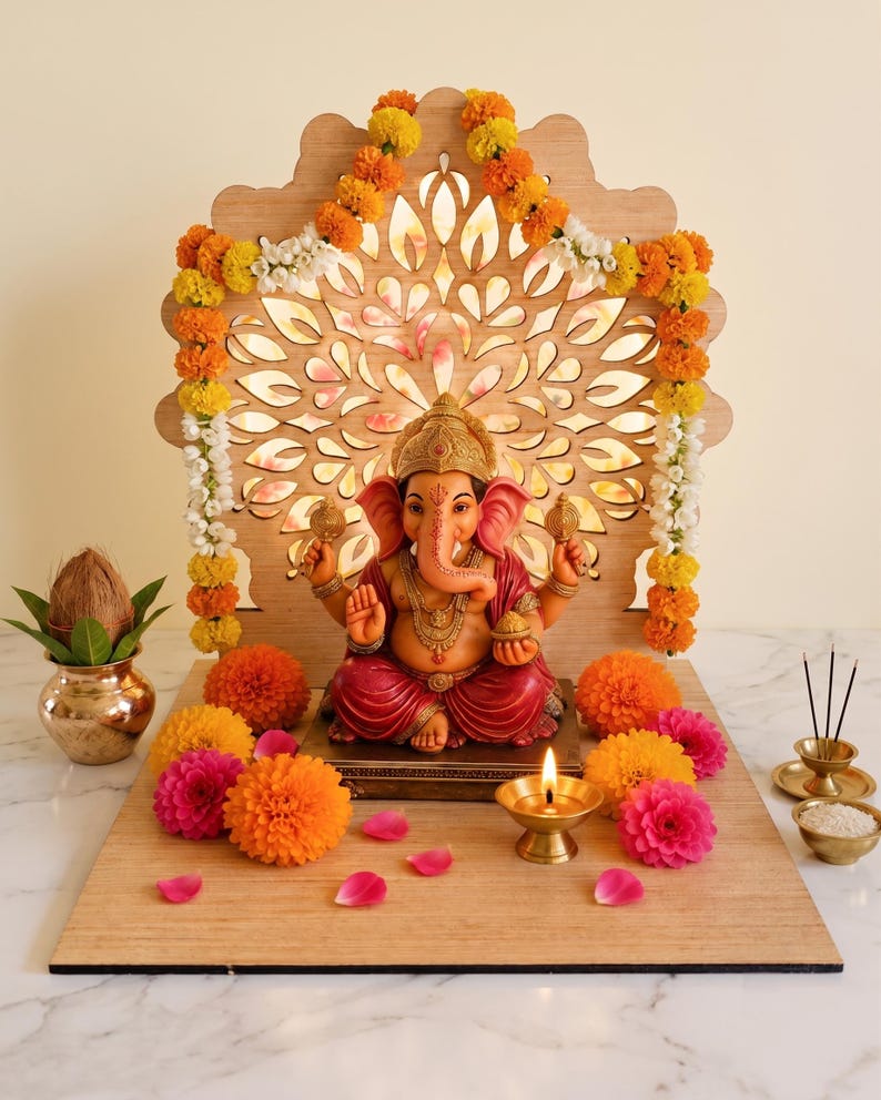 Op de afbeelding: Een Ganesha-beeld met een roze huid en rode kleding staat op een houten voetstuk voor een lasergesneden houten achtergrond. Het geheel is versierd met oranje en roze afrikaantjes, een koperen olielamp, een kokosnoot in een koperen pot en wierook.
