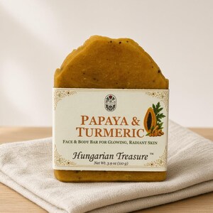 May include: A mustard-yellow rectangular face and body soap bar labeled Papaya & Turmeric Hungarian Treasure, featuring a graphic of a papaya and turmeric root, with a net weight of 3.9 oz printed on the white paper wrapper.