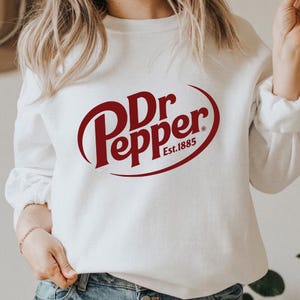 May include: White sweatshirt featuring the Dr Pepper logo in a maroon color. The logo is in a stylized font within an oval shape, with "Est.1885" below. The sweatshirt is a casual style.