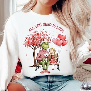 Op de afbeelding: Witte sweatshirt met de roze tekst "ALL YOU NEED IS LOVE". De afbeelding toont een groen personage, een meisje en een hond die op een bank zitten onder een hartvormige boom. Roze hartballonnen zweven in de lucht.