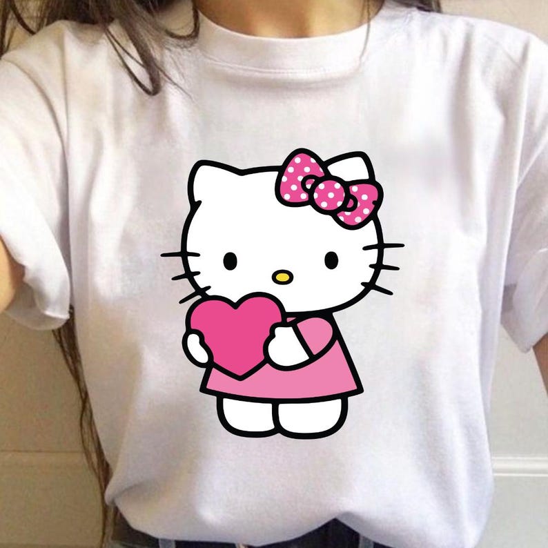 Puede incluir: Camiseta blanca con una ilustraci&oacute;n de dibujos animados de Hello Kitty sosteniendo un coraz&oacute;n rosa. Hello Kitty lleva un vestido rosa y un lazo rosa con lunares blancos. El dise&ntilde;o tiene contornos negros.