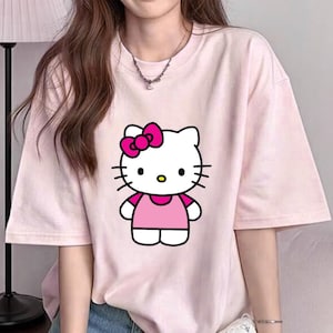 Puede incluir: Camiseta rosa claro con un dise&ntilde;o de dibujos animados de Hello Kitty. El gr&aacute;fico muestra a Hello Kitty con un lazo rosa, un vestido rosa y bigotes negros. La camiseta tiene mangas cortas y cuello redondo.