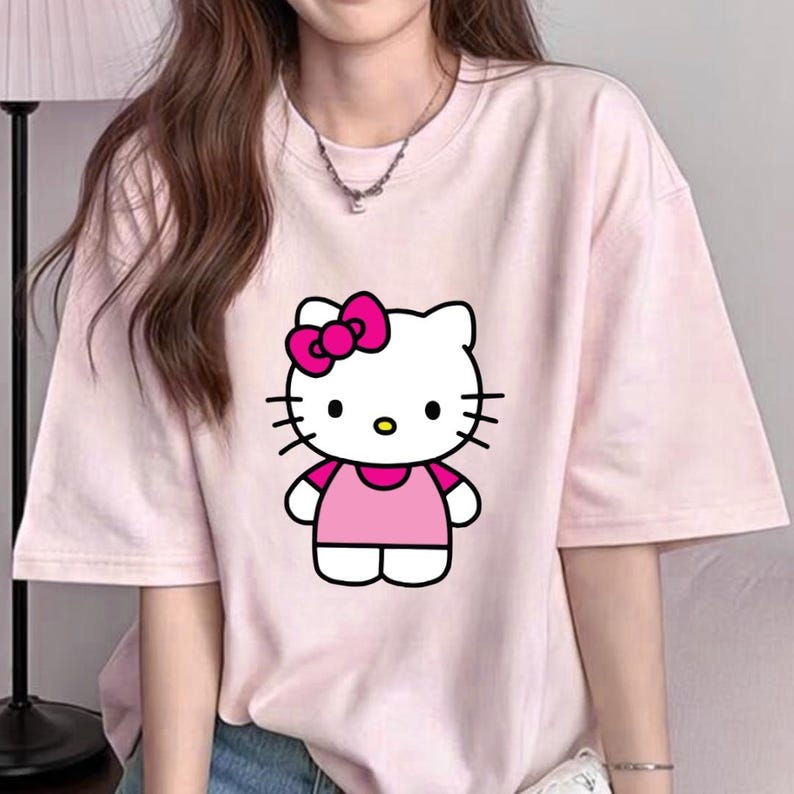 Puede incluir: Camiseta rosa claro con un dise&ntilde;o de dibujos animados de Hello Kitty. El gr&aacute;fico muestra a Hello Kitty con un lazo rosa, un vestido rosa y bigotes negros. La camiseta tiene mangas cortas y cuello redondo.