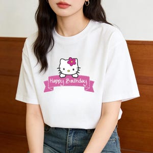 Puede incluir: Camiseta blanca con un dibujo de un gato de dibujos animados, una flor rosa y el texto "Happy Birthday" en una pancarta rosa. La camiseta tiene mangas cortas y cuello redondo.