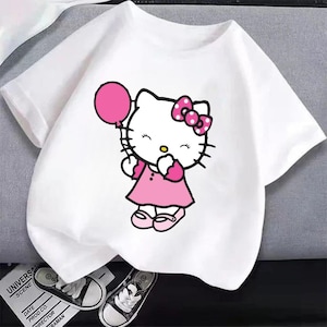 Puede incluir: Camiseta blanca con una ilustraci&oacute;n de dibujos animados de Hello Kitty sosteniendo un globo rosa. Hello Kitty lleva un vestido y un lazo rosas. Zapatillas deportivas en blanco y negro visibles en la parte inferior.
