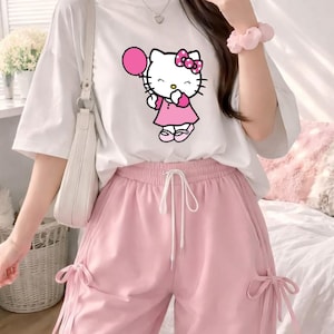 Puede incluir: Camiseta blanca con un gr&aacute;fico de Hello Kitty sosteniendo un globo rosa, combinada con pantalones rosas con cord&oacute;n. El conjunto incluye un bolso de hombro blanco. La est&eacute;tica general es suave y femenina, con un enfoque en el personaje de dibujos animados.