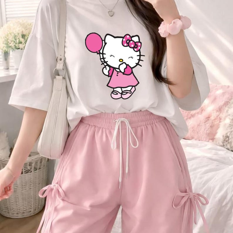 Puede incluir: Camiseta blanca con un gr&aacute;fico de Hello Kitty sosteniendo un globo rosa, combinada con pantalones rosas con cord&oacute;n. El conjunto incluye un bolso de hombro blanco. La est&eacute;tica general es suave y femenina, con un enfoque en el personaje de dibujos animados.