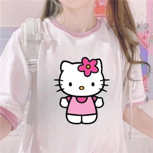 Puede incluir: Camiseta blanca con ribete rosa con un gr&aacute;fico de Hello Kitty. El gr&aacute;fico muestra a Hello Kitty con un vestido rosa y una flor rosa en la cabeza. La camiseta tiene mangas cortas y cuello redondo.