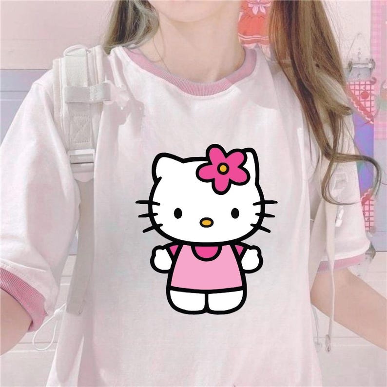 Puede incluir: Camiseta blanca con ribete rosa con un gr&aacute;fico de Hello Kitty. El gr&aacute;fico muestra a Hello Kitty con un vestido rosa y una flor rosa en la cabeza. La camiseta tiene mangas cortas y cuello redondo.