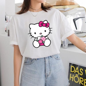 Puede incluir: Camiseta blanca con un gr&aacute;fico de Hello Kitty sosteniendo flores. El personaje de dibujos animados tiene un lazo rosa y est&aacute; delineado en negro. La camiseta se combina con pantalones cortos de mezclilla.