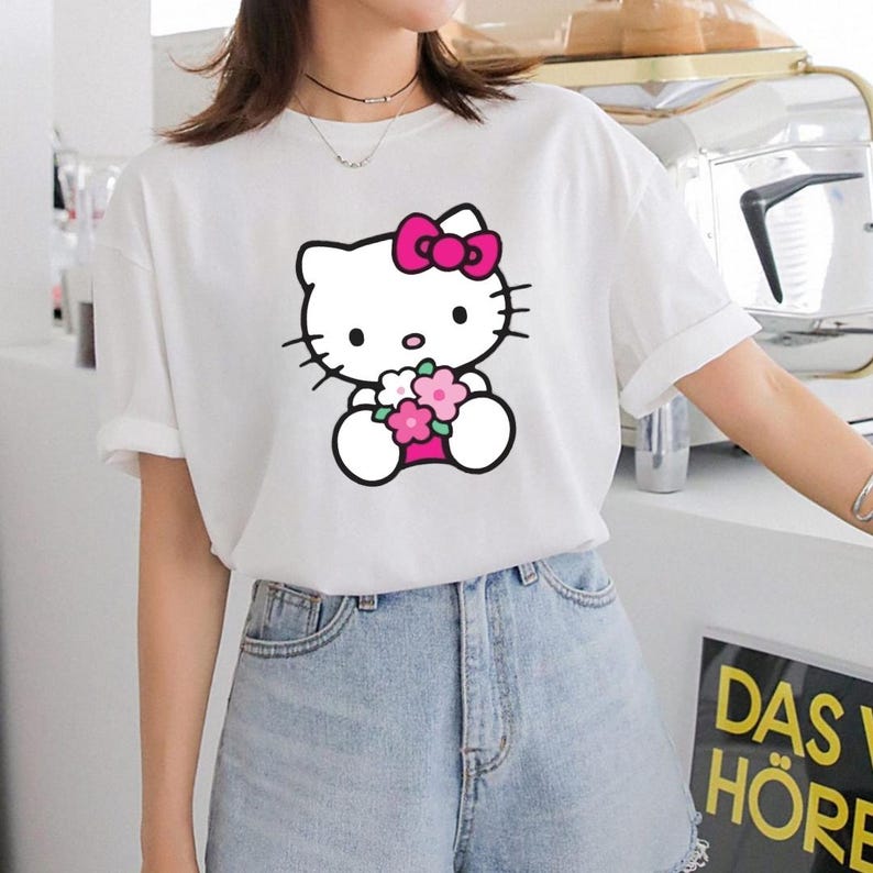 Puede incluir: Camiseta blanca con un gr&aacute;fico de Hello Kitty sosteniendo flores. El personaje de dibujos animados tiene un lazo rosa y est&aacute; delineado en negro. La camiseta se combina con pantalones cortos de mezclilla.