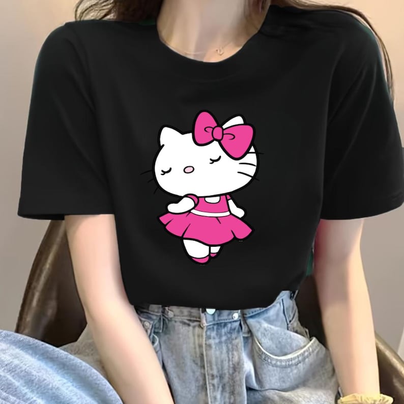 Puede incluir: Camiseta negra con un gr&aacute;fico de Hello Kitty con vestido y lazo rosa. El personaje de dibujos animados tiene los ojos cerrados y la cara blanca. La camiseta es de cuello redondo.