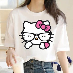 Puede incluir: Camiseta blanca con un gr&aacute;fico de Hello Kitty con gafas y un lazo rosa. El personaje de dibujos animados est&aacute; delineado en negro con un lazo rosa y un objeto en forma de coraz&oacute;n rosa. La camiseta tiene mangas cortas.