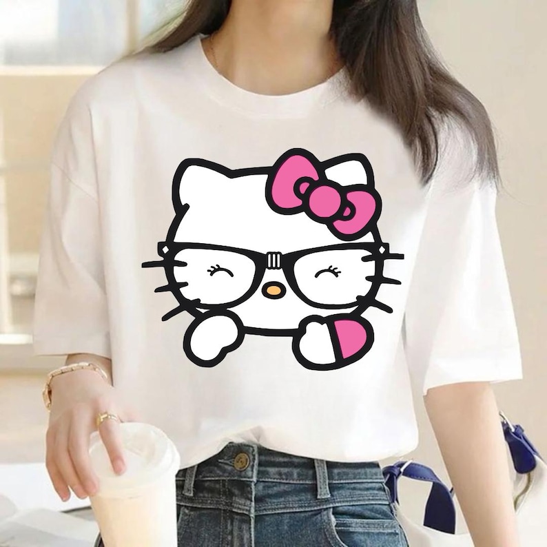 Puede incluir: Camiseta blanca con un gr&aacute;fico de Hello Kitty con gafas y un lazo rosa. El personaje de dibujos animados est&aacute; delineado en negro con un lazo rosa y un objeto en forma de coraz&oacute;n rosa. La camiseta tiene mangas cortas.