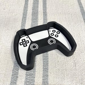 May include: A black and white controller-shaped tray. The tray features a design of a video game controller, with white buttons and a black frame. The tray is designed to hold small items and is placed on a striped surface.