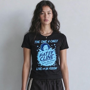 May include: A black short-sleeved t-shirt featuring a light blue graphic print. The design includes the text THE ONE & ONLY PATSY CLINE, Walkin After Midnight, Sweet Dreams, and LIVE and IN PERSON, along with a stylized portrait of the singer.
