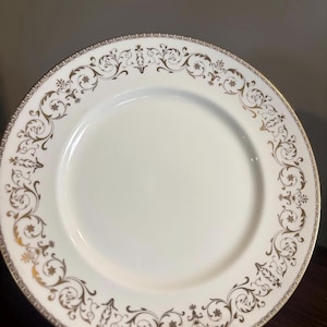 Puede incluir: Un plato de cerámica blanca con un diseño floral dorado en el borde. El plato presenta un delicado borde dorado y un intrincado diseño de espirales y motivos florales. El plato se presenta sobre un soporte de metal plateado.