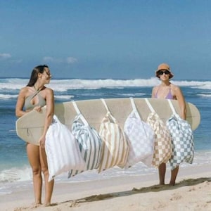 Puede incluir: Una tabla de surf con seis bolsas con cordón colgando. Las bolsas tienen varios patrones, incluyendo rayas y diseños a cuadros. Dos personas están de pie en una playa arenosa, sosteniendo la tabla de surf. El océano y el cielo azul están en el fondo.