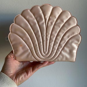 May include: A light pink, shell-shaped clutch with a scalloped edge. The clutch has a quilted design with vertical lines radiating from the center. The clutch is held in a person's hand.