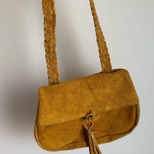 May include: A mustard yellow suede crossbody bag with a braided leather strap and a tassel.