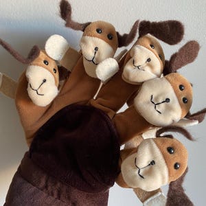 May include: A hand puppet featuring five dog-shaped finger puppets. Each puppet has a brown and tan head with floppy ears, black eyes, and a stitched smile. The puppet's body is a dark brown color.