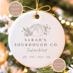 May include: A white ceramic ornament with a gold string for hanging. The ornament features a line drawing of a loaf of bread surrounded by floral elements. Text includes "SARAH'S SOURDOUGH CO." and "Fresh from the heart".