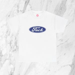 Pode incluir: T-shirt branca com um logótipo oval azul que diz "Fuck" numa fonte estilizada. A t-shirt é feita de um material macio e tem uma gola redonda.