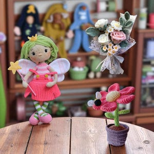 Puede incluir: Una colección de artículos de ganchillo hechos a mano, que incluyen una muñeca de hadas con cabello verde y un vestido rosa, un ramo de flores y una flor roja con una abeja. Los artículos se exhiben sobre una superficie de madera.