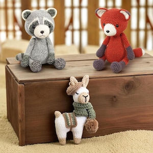 Op de afbeelding: Drie handgemaakte gehaakte knuffels: een grijze wasbeer, een rode vos en een witte lama. De wasbeer heeft een zwart masker en een grijze staart. De vos heeft paarse accenten. De lama draagt een groene sjaal en een bruine hoed.