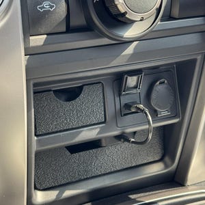 May include: Close-up of a car's dashboard featuring a black console with a small drawer, a USB port with a charging cable, and a 12V power outlet. The A/C controls and other buttons are visible.