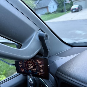 May include: A digital display screen mounted on a vehicle's dashboard, showing various numerical data in red, blue, and white. The screen is attached to a black bracket, and the vehicle's interior is visible.