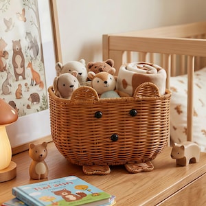May include: A brown wicker basket shaped like a bear, filled with stuffed animals and a rolled blanket. The basket has black button eyes and feet. A wooden bear figurine and a children's book are on the wooden surface.