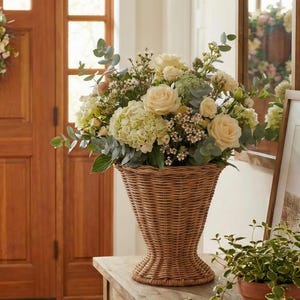 May include: A large floral arrangement in a woven wicker basket. The bouquet features cream-colored roses, hydrangeas, and various greenery. The basket is a light brown color and sits on a wooden table.